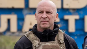 Лисий чоловік у камуфляжному тактичному жилеті перед синьою вивіскою