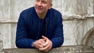 Чоловік у синій стьобаній куртці зі схрещеними руками спирається на камяний парапет