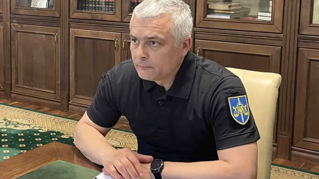 Чоловік із сивим волоссям у чорному поло з нашивкою сидить за столом в кабінеті