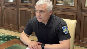 Чоловік із сивим волоссям у чорному поло з нашивкою сидить за столом в кабінеті